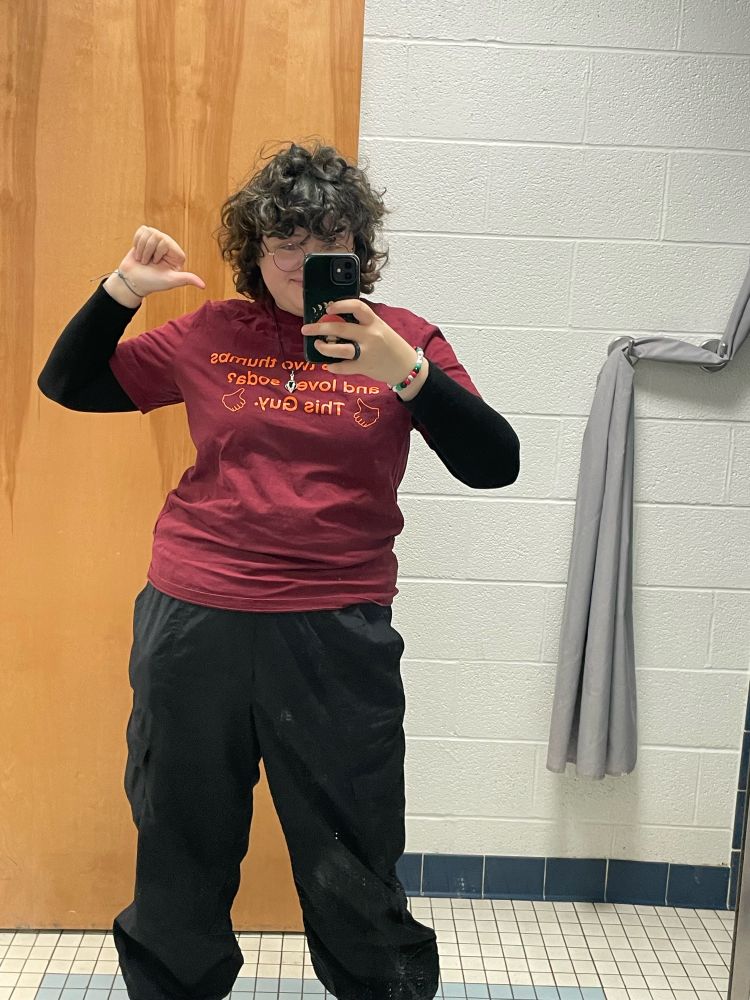 person with dark hair wearing a maroon tshirt with orange text saying “who loves soda? this guy.” and two orange thumbs pointing towards the top of the shirt. person is wearing dark pants and a black turtle neck underneath while using their thumb to point at themselves