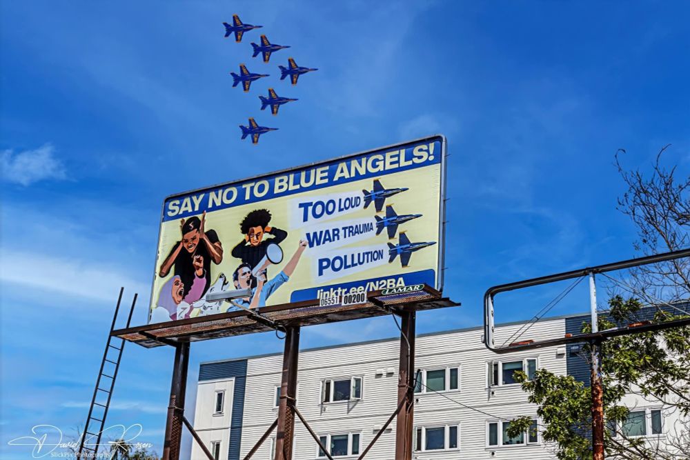 Six 'blue angels' warplanes flying in tight formation over the billboard with the banner "say no to blue angels"
Photo by David Rosen / SlickPix