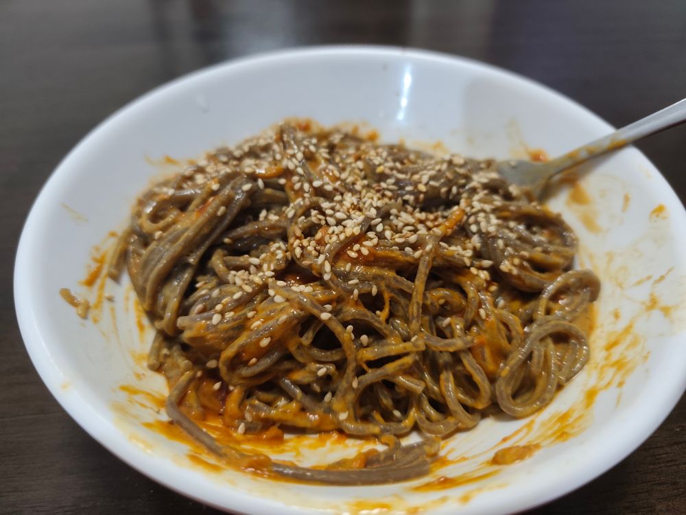 A bowl of brown soba noodles in a peanut sauce, sprinkled with sesame seeds.