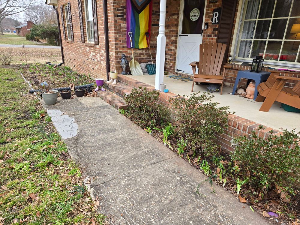 A picture of greenish grass, grey concrete sidewalk, cleaned out flower beds, a brick house, Adirondack chairs, and a Pride flag.
