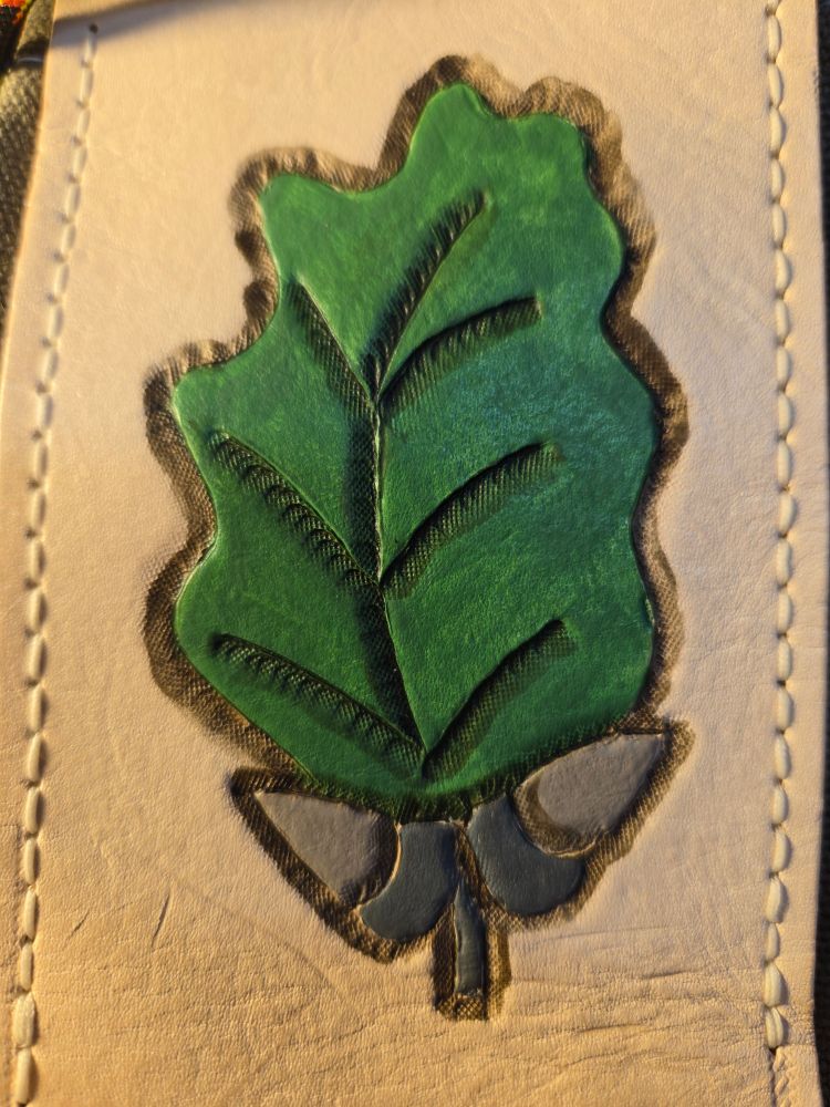 Tanned leather stitched to a canvas bag. The leather is light tan and has a leaf and two acorns carved and stamped into it. The leaf is painted green and the acorns are grey while the design has darker shades around to imply shadow.