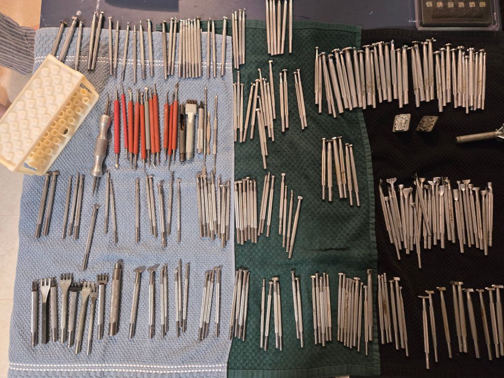 An image of leatherworking stamps laid out on blue, green, and black towels all on a blue folding table. The tools are all shiny silver and a few have red handles. There is a white plastic stand to the side of the tools.