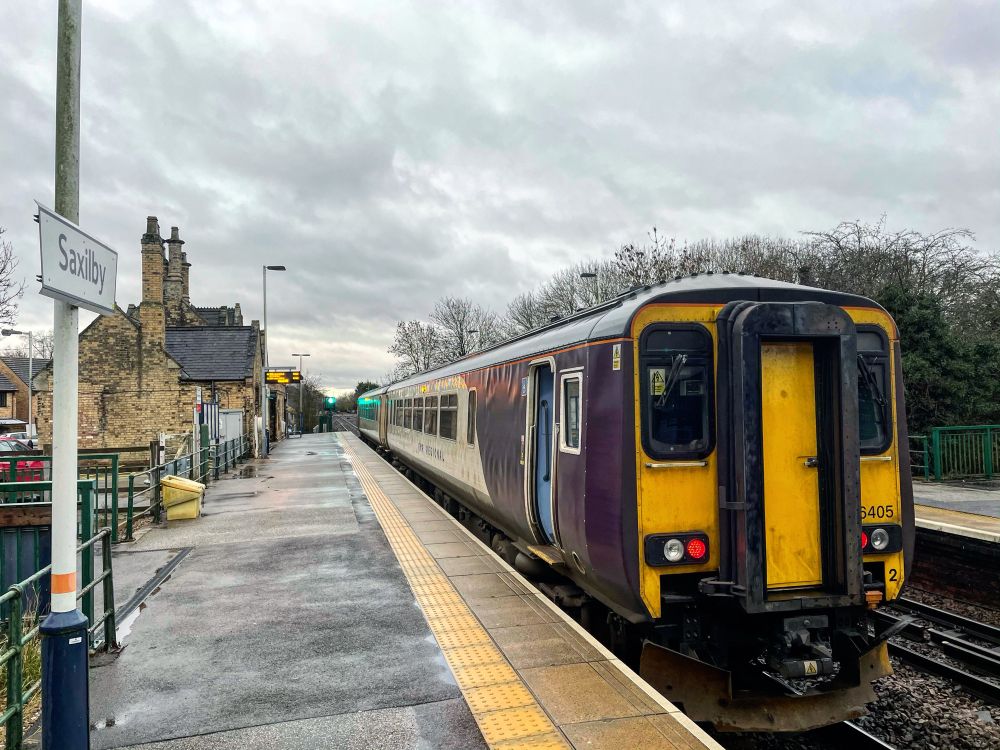An empty, damp platform at Saxilby with the starting signal at green, grey skies and leafless trees. The station building with its multiple chimneys can be seen to the left. The train is EMR purple, white and grey liveried 156405 with yellow end warning panel. The train is heading away from the photographer so tail lights are lit. There are no people visible.