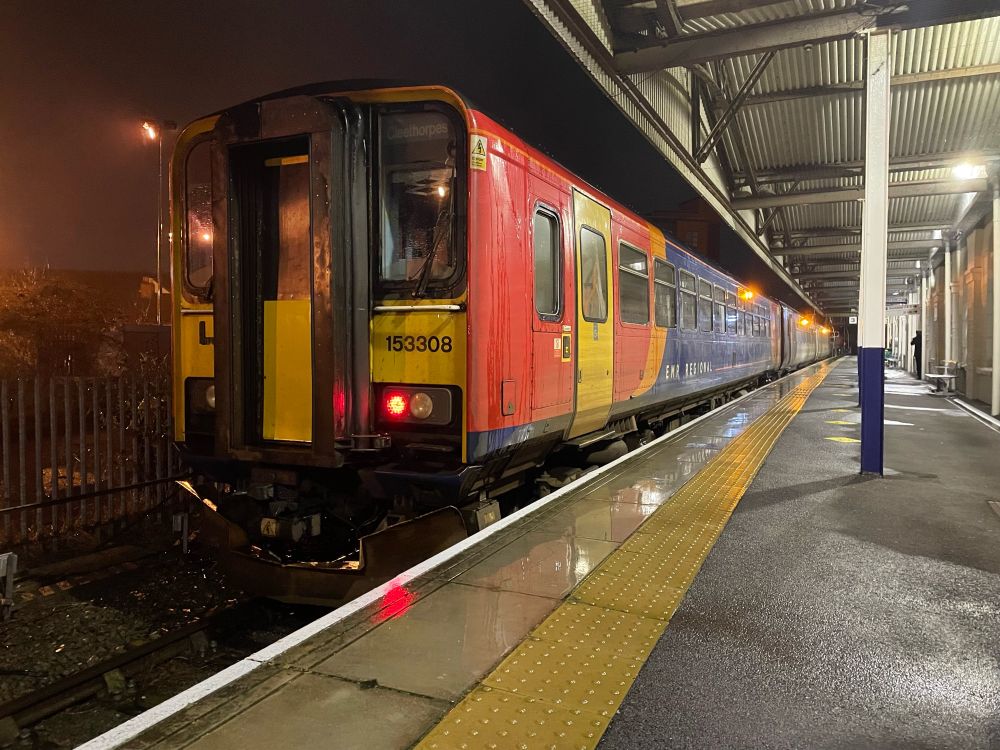 153308 in blue, red and orange East Midlands trains livery with EMR regional titles in bay platform 3 at Grimsby. A 156 in EMR interim purple, gray and white livery is at the front. It is dark and the station lights are lit and you can see drizzle in the air. Rain standing on the platform edge is reflecting the lights.