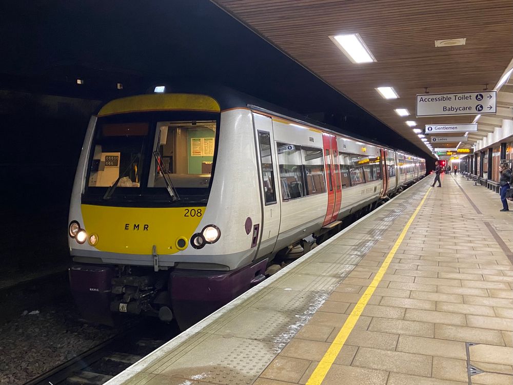 170208 in white Anglia livery with red doors and grey skirt, along with some purple skirts. The headlights are on and the cab light is on. People are walking around on the platform and the rear door on the front coach is partially opening. It is dark and the station lights are on.