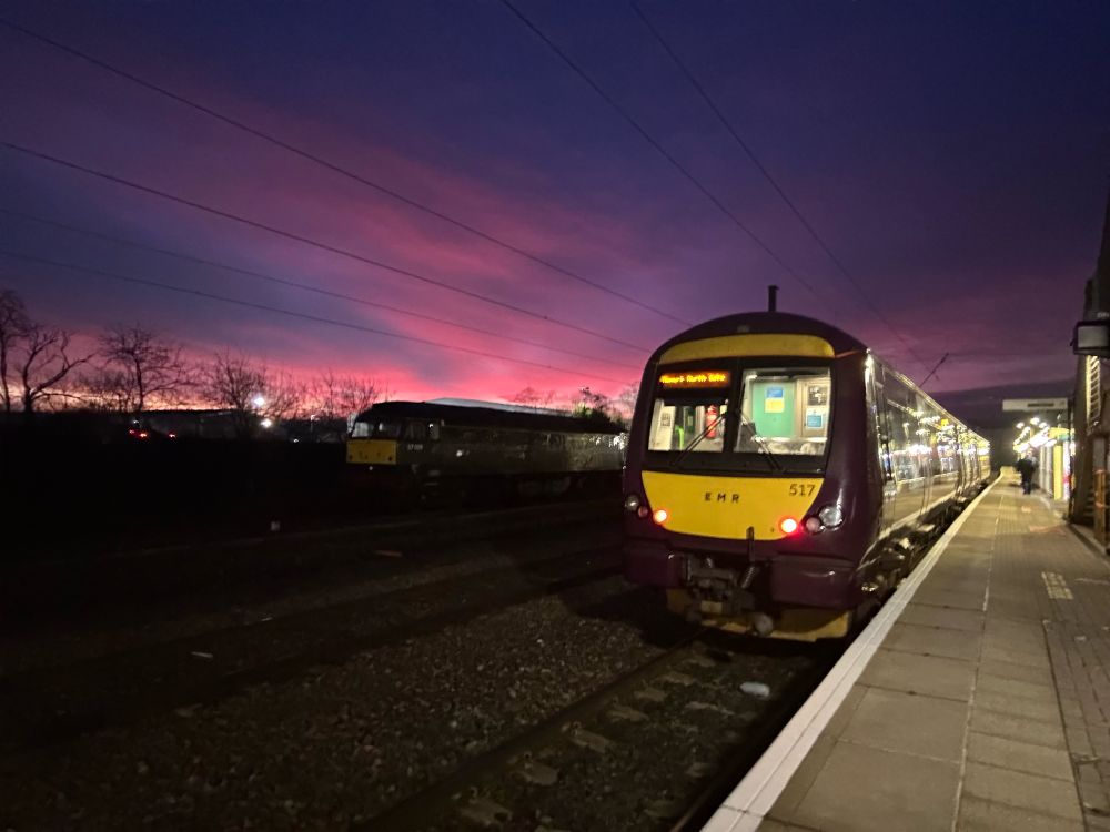 170517 in EMr purple livery with tail lights illuminated on platform 3 at Newark north gate. There is a green West Coast 57 behind in the dark. The sky above is pink and purple with the orange of the sunrise to the bottom.