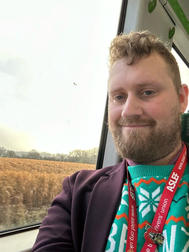 Me wearing my purple EMR blazer, a turquoise and orange Beanz Meanz Crimbo jumper and ASLEF lanyard with SWR class 442 poppy badge. Out the window you can see fields, trees and gray skies.