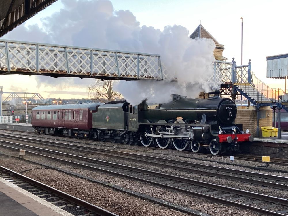 Steam loco 45596 ‘Bahamas’ in steam at Lincoln station with the footbridge above. The locomotive is green and pulling a crimson coach.