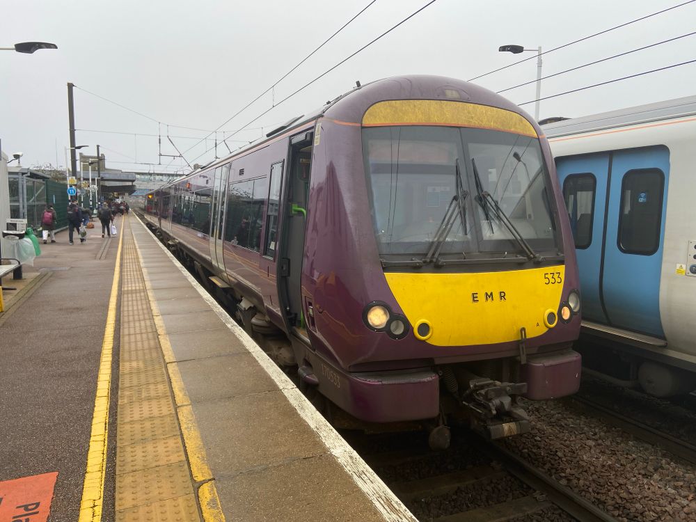 170533 in EMR purple livery in platform 1
At Peterborough. The platform is busy with people behind the yellow line and tactile paving. You can see a Thameslink grey class 700 with blue doors in the adjacent platform. The sky is also grey and overcast.
