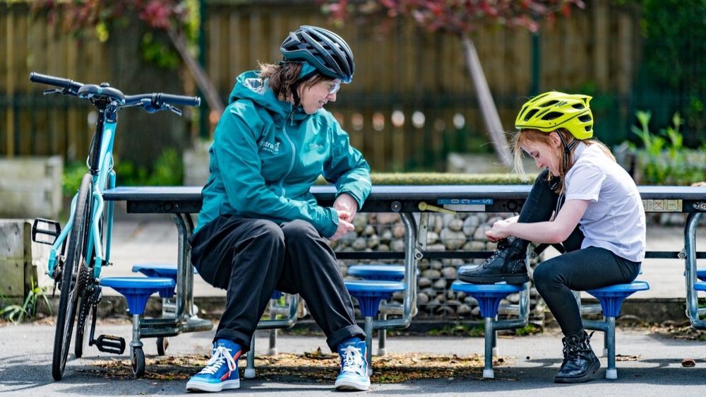 Sustrans I Bike Officer sitting with a primary school pupil who has stopped to tie their shoelaces during a cycling skills session in the playground.