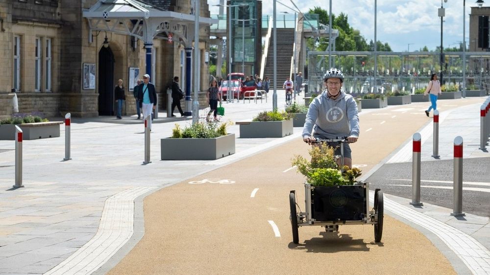 Person cycling a cargo bike carrying plants, on a segregated cycle lane outside a train station