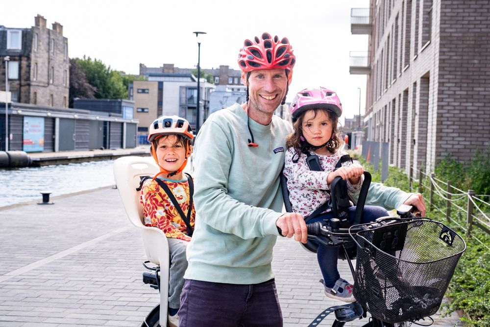 A dad and his two children standing with their bike on the newly improved canal towpath in Fountainbridge, Edinburgh. All three ride together on the same bike and are wearing helmets.