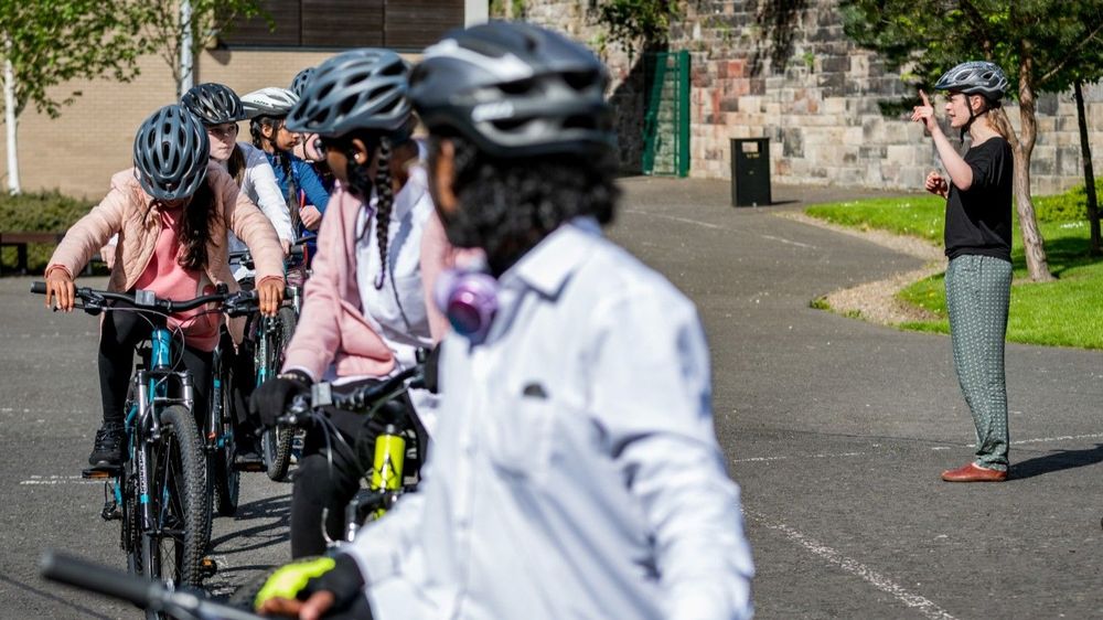 Teenage girls lined up on bikes, learning cycling skills in a school playground during an I Bike session.