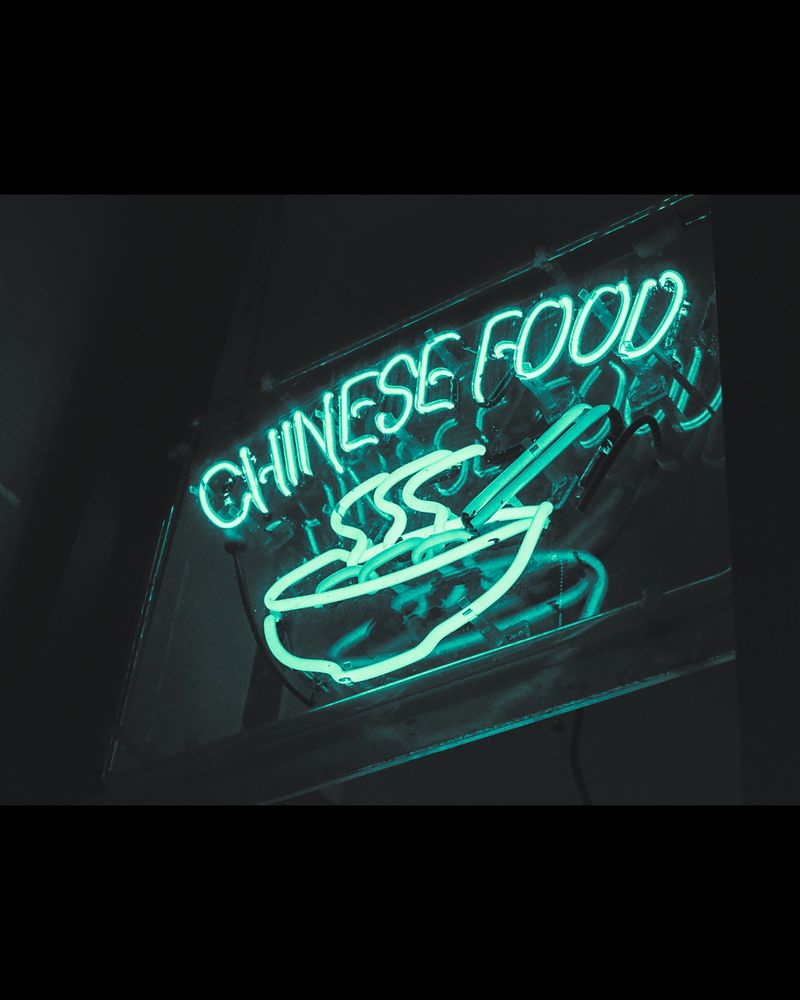 Black and white photo of a neon sign that says, "Chinese food" and depicts a steaming bowl with chopsticks. The photo has been edited to make the sign appear green.