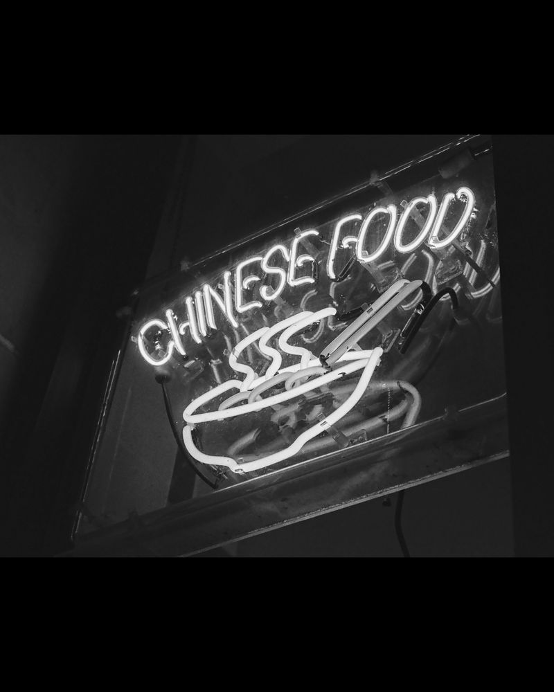 Black and white photo of a neon sign that says, "Chinese food" and depicts a steaming bowl with chopsticks.