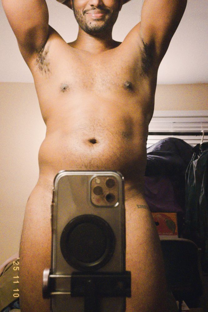 a nude figure is standing in front of a mirror, their arms up behind their head, their phone obscuring their crotch. they’re smiling.