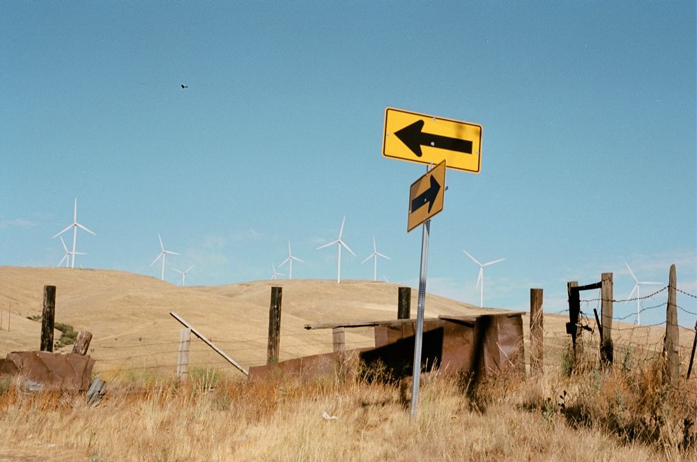 A yellow arrow signpost that is found on a sharp road corner. Rolling hills covered in dry grass, with wind turbines in the background.