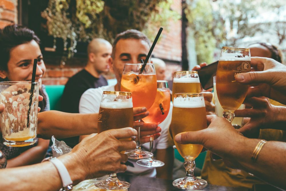 Menschen im Sommer draußen in einer Bar, die sich mit Bier und anderen alkoholischen Getränken zuprosten. Foto: Unsplash/Fred Moon