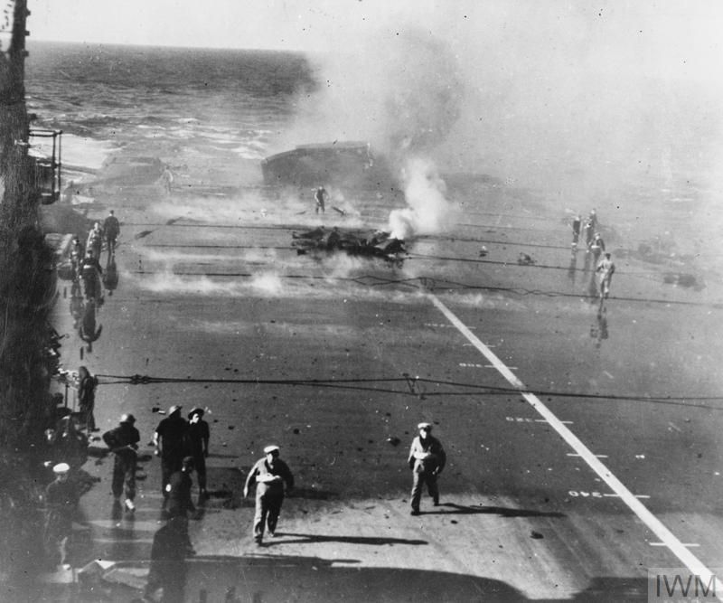 HMS Illustrious after a bomb hit on the aft section of the flight deck. A large hole is pictured, with smoke coming from the rear of the ship. Sailors are seen on the flight deck attending to the damage. 
