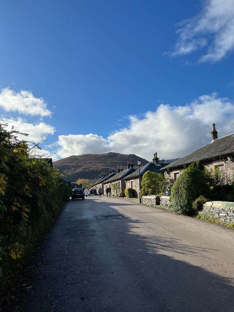 Main Street of Luss looking at hills to the west. Stone houses and low stone walls. Blue sky with clouds. 