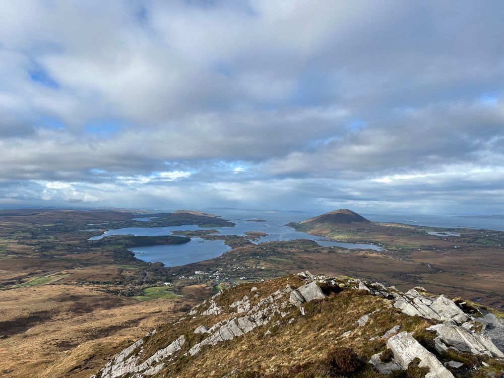 Higher up view of the small bay at Letterfrack that opens onto the Atlantic Ocean. Rocky knoll in foreground. Heather and bog below peak. Clouds with some blue sky