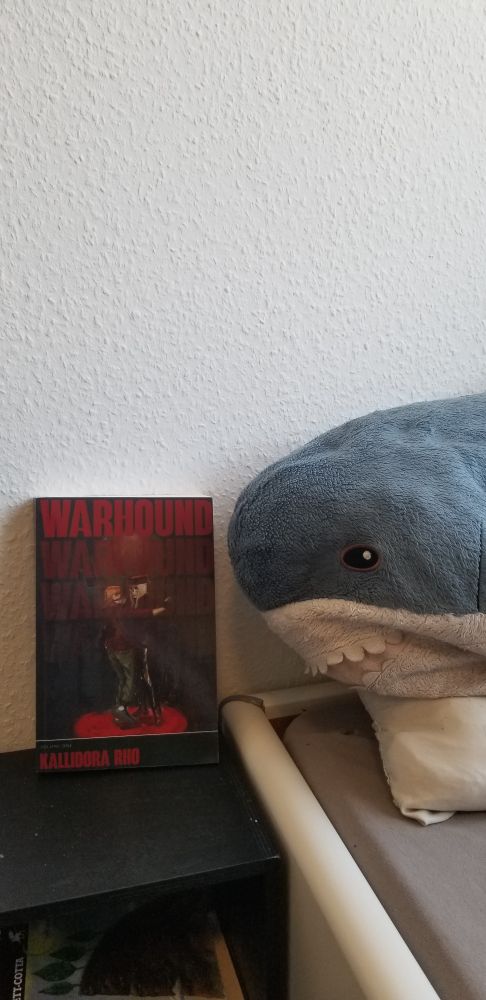 A copy of Warhound by Kallidora Rho propped up on my bedside table with my blåhaj next to it.