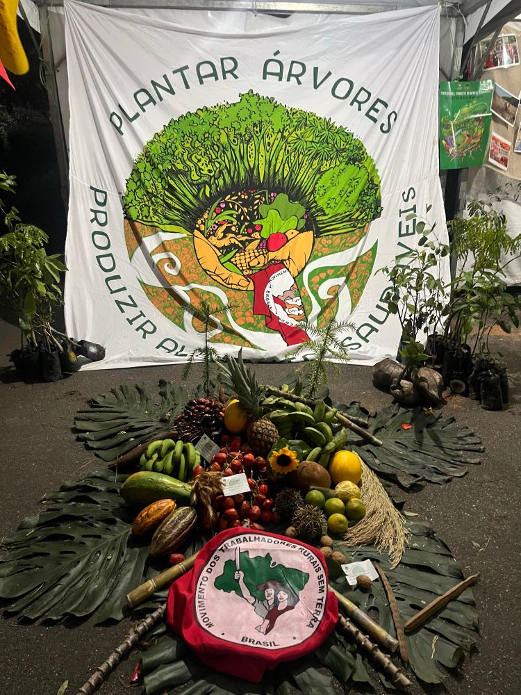 Alimentos no chão em cima de folhas de bananeira com uma bandeira do mst dobrada na frente, ao fundo uma bandeira maior, onde está escrito "plantar árvores" seguido de "produzir alimentos saudáveis" abaixo. No meio uma ilustração do campo, com árvores, alimentos, e mãos segurando a bandeira do mst. Nos cantos mudinhas de plantas. 