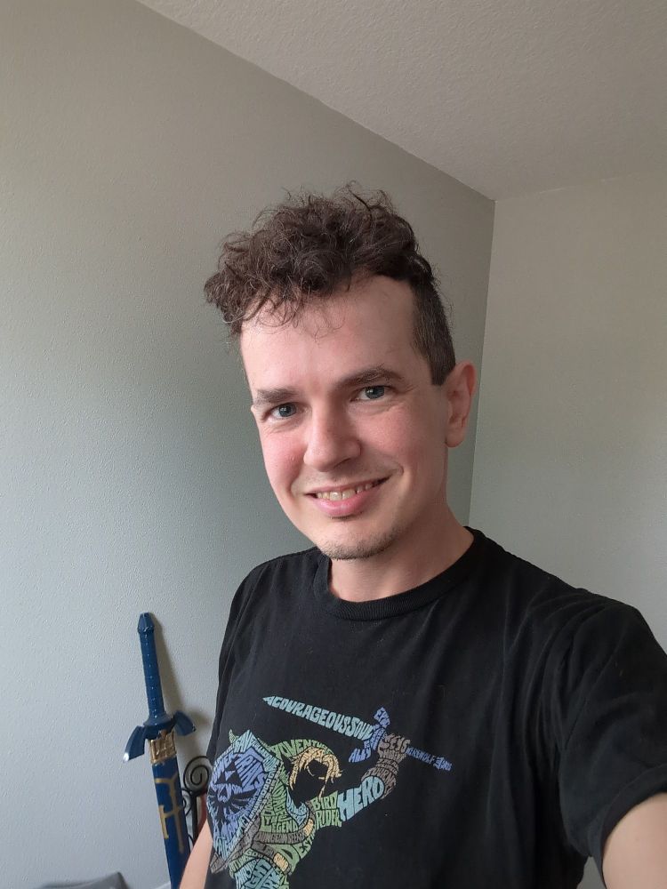 A selfie of me smiling at the camera while wearing a black Zelda T-shirt. Meanwhile a replica master sword can be seen leaning against the wall behind me.
