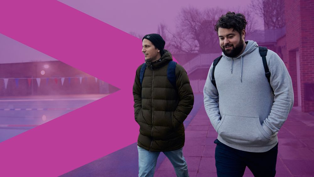 Two young men walk side by side near an outdoor swimming pool on a chilly day, both wearing backpacks and warm jackets. There is a purple GM Moving arrow behind them.