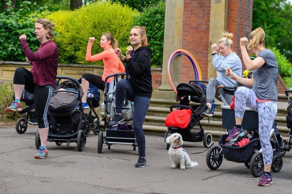 A group of women exercising outdoors while standing next to prams. They are raising knees and fists in a fitness routine.