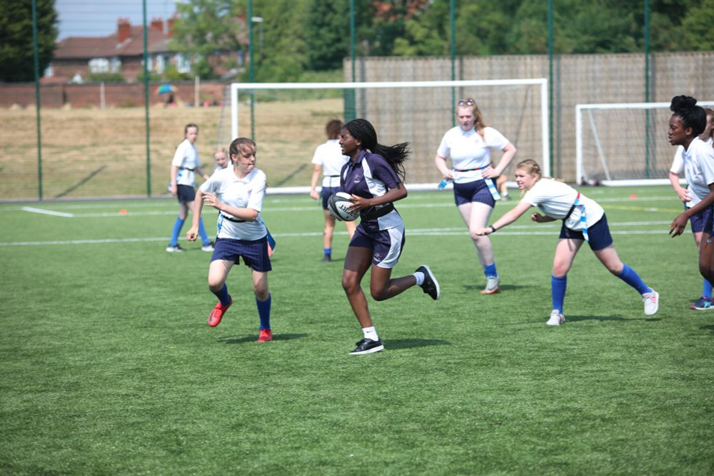A group of young women and girls play rugby outside on a sunny day, while wearing sports gear.