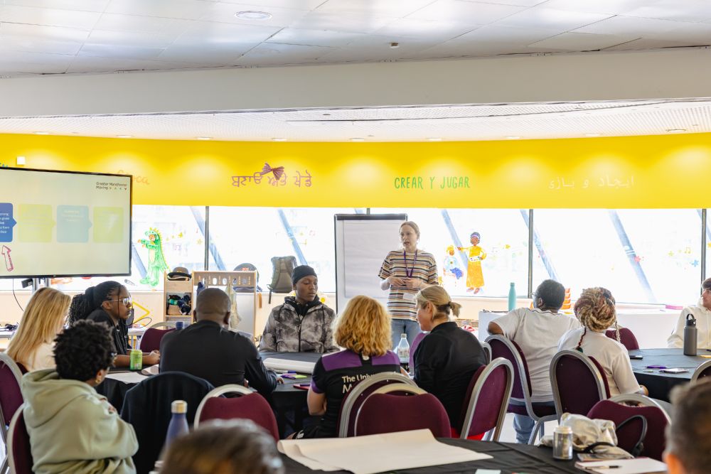 A group workshop with one person presenting at the front, while others listen while seated.