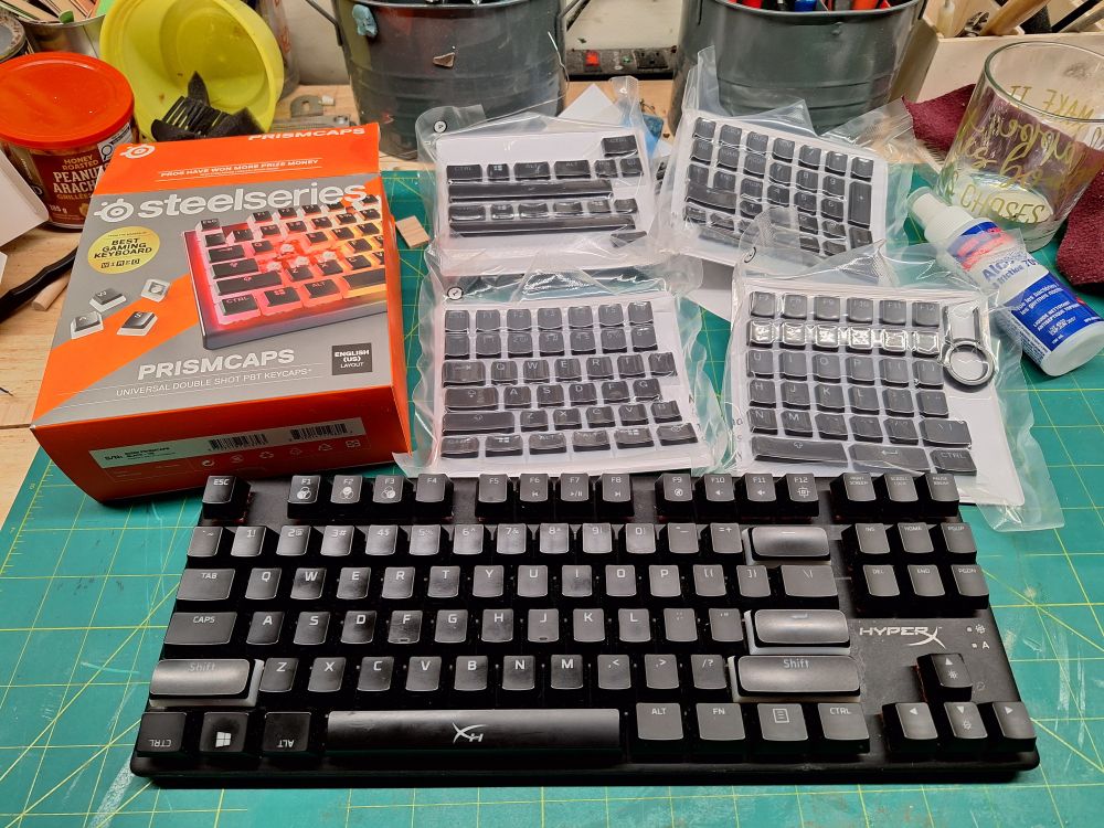An HyperX TKL mechanical keyboard on a workbench, with a box of SteelSeries Prismcaps laid out behind it.