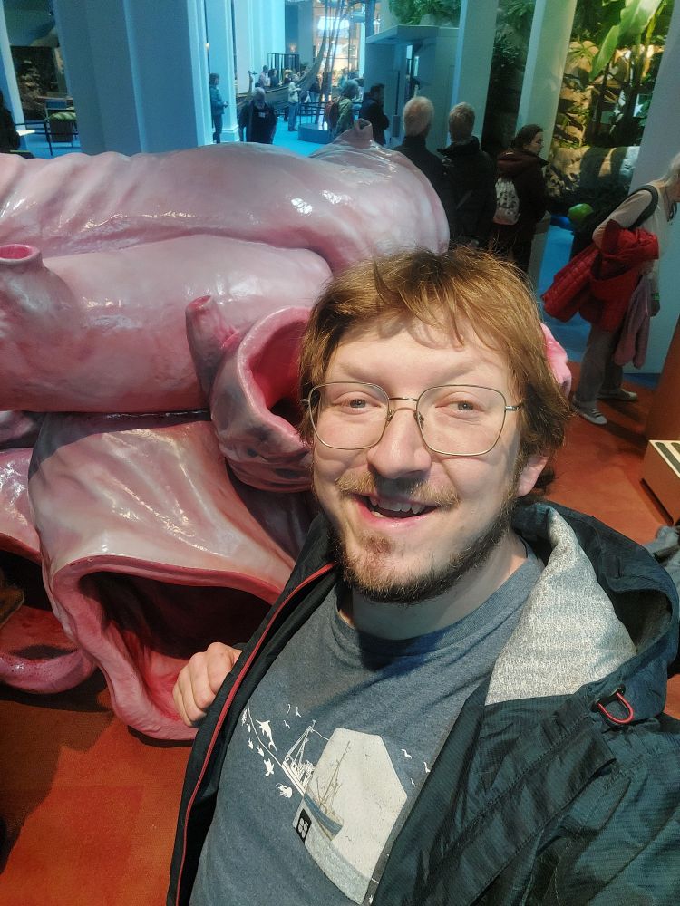 Selfie of me, a mid thirties male with glases and blue clothing. I am standing in a museum in front of a model of a blue whale heart, wich is as tall as I am.