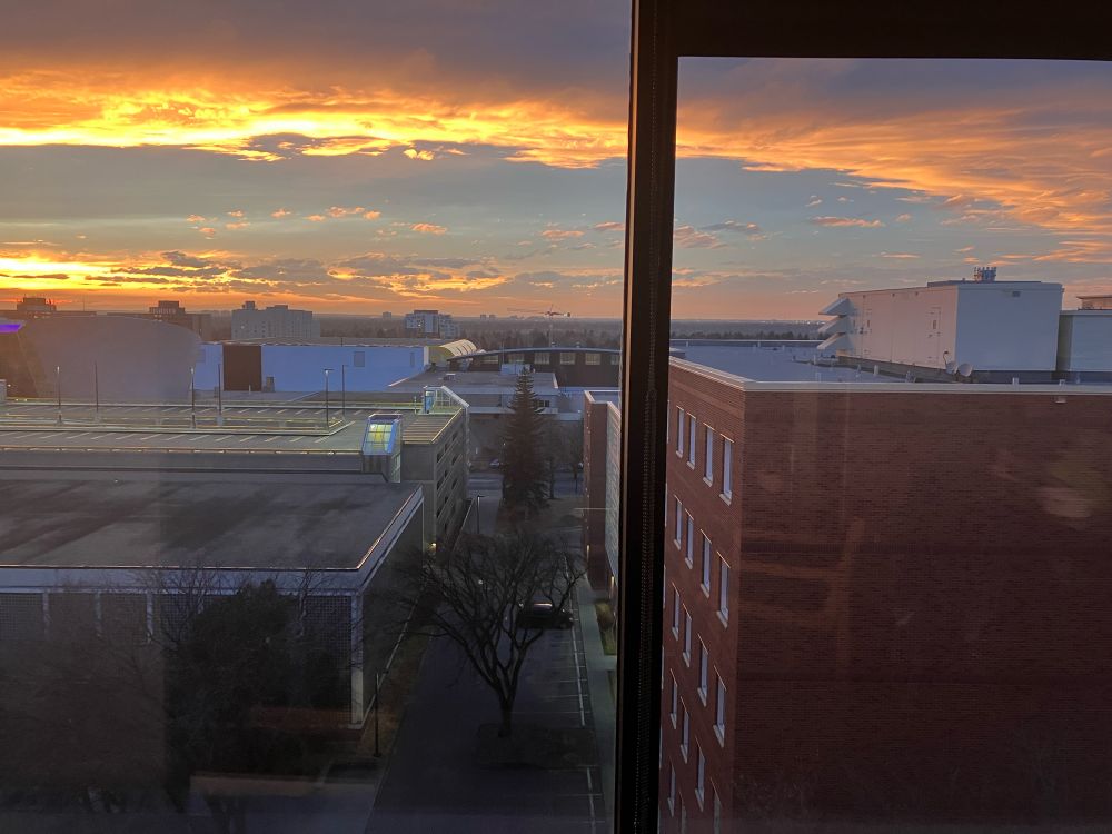 A photo from an upper-level office window with other buildings in the foreground and a sunset in the background. Two parallel horizontal banks of clouds are lit with blazing golden hues. 