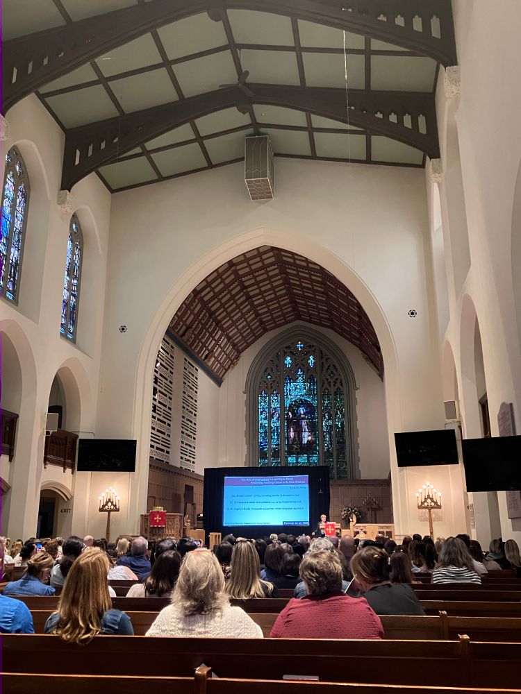 Dr. Jamie Metsala stands beside a screen, giving a keynote lecture under a massive stained glass window in a church at ResearchEd in Toronto. 