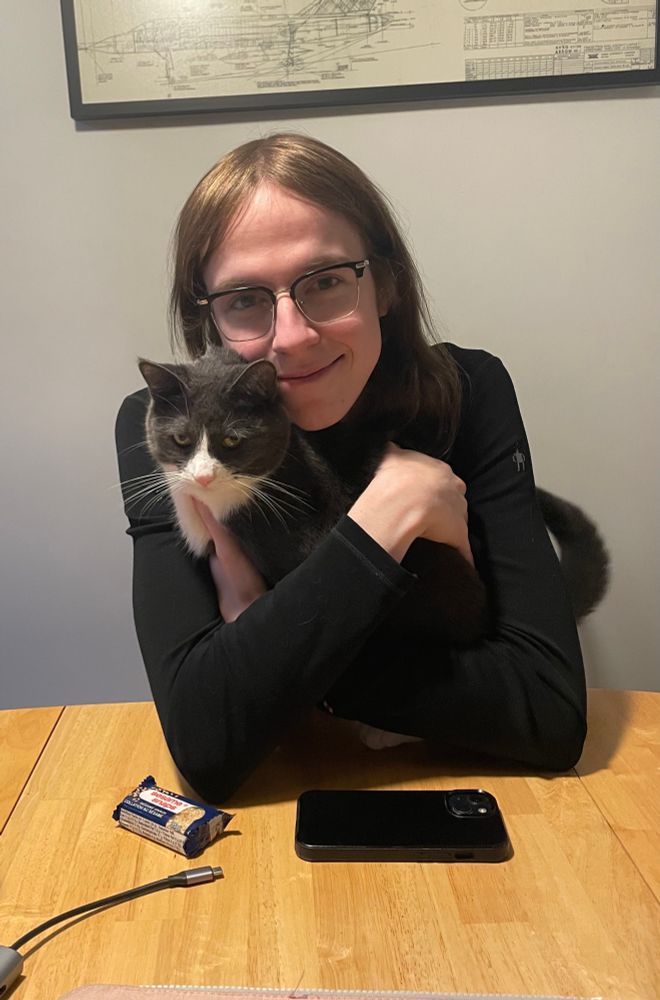 a photo of sam, a trans woman, sitting at a table and hugging jacques, a grey cat. She is looking at the camera and smiling, and is wearing a black long sleeve. 