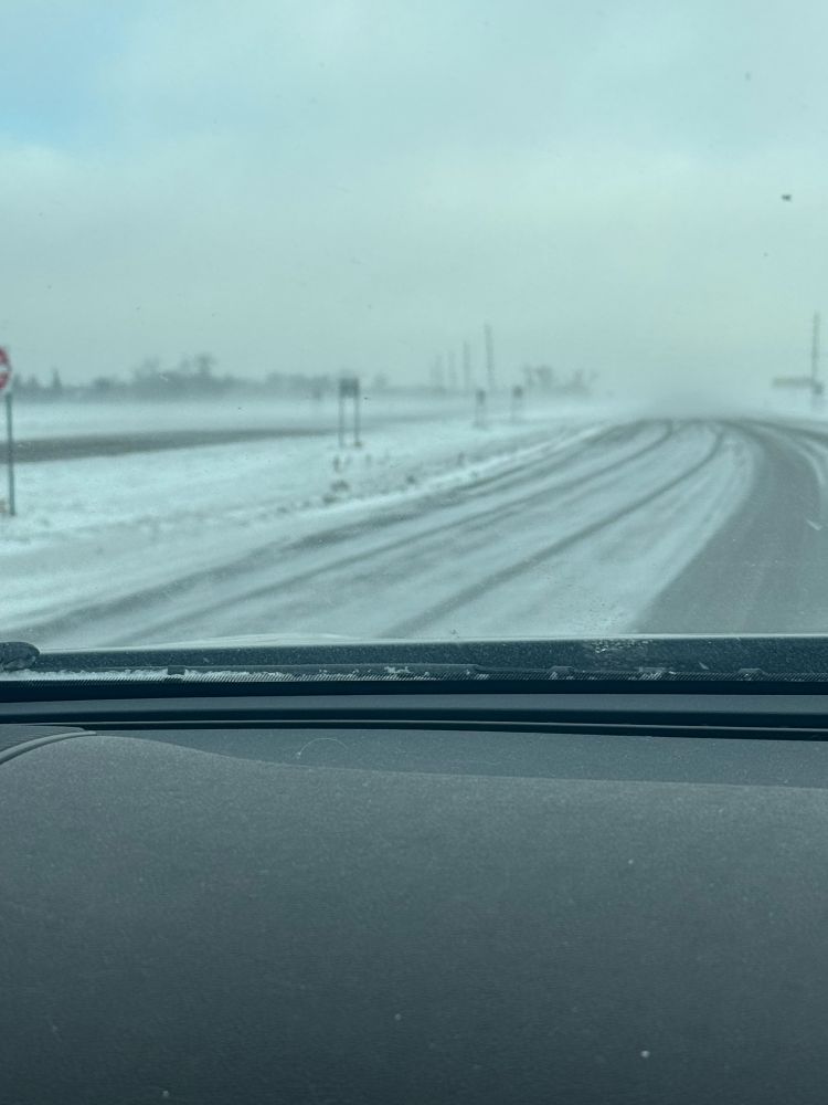Photo through car windshield of rural highway covered with blowing snow under a dreary, gray, windy sky