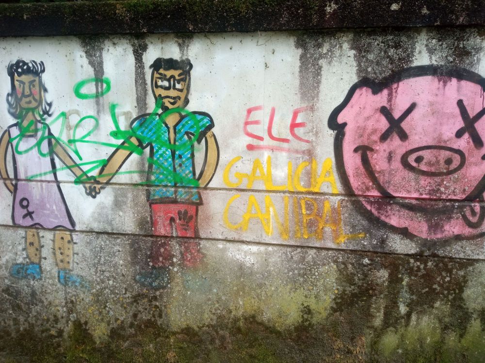 Foto dun grafiti nun muro. De esquerda a dereita podemos ver:

Unha parella visiblemente queer dándose a man. Unha é unha persoa peluda con bigote e un vestido rosa co simboliño da muller. A outra leva unha camisa azul e uns pantalóns de chándal vermellos. 

Un texto no que pon, en vermello, "ELE", e debaixo en amarelo "GALICIA CANÍBAL".

Á dereita de todo hai un debuxo da cara dun cocho cos ollos en X e a lingua de fóra que recorda un pouco ao logotipo de Nirvana.