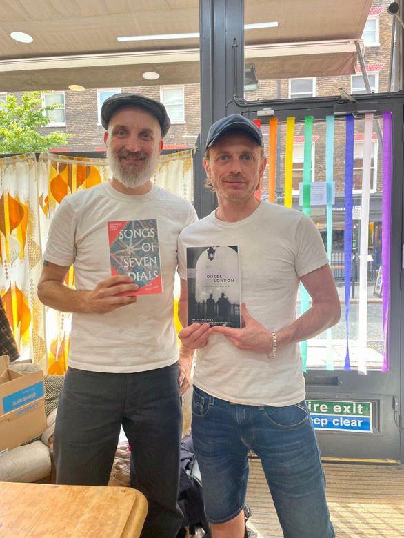 A middle aged man with a white beard wearing a white t-shirt and jeans looks awkward holding a copy of his book Songs of Seven Dials: An intimate history of 1920s and 1930s London in a bookshop in London.