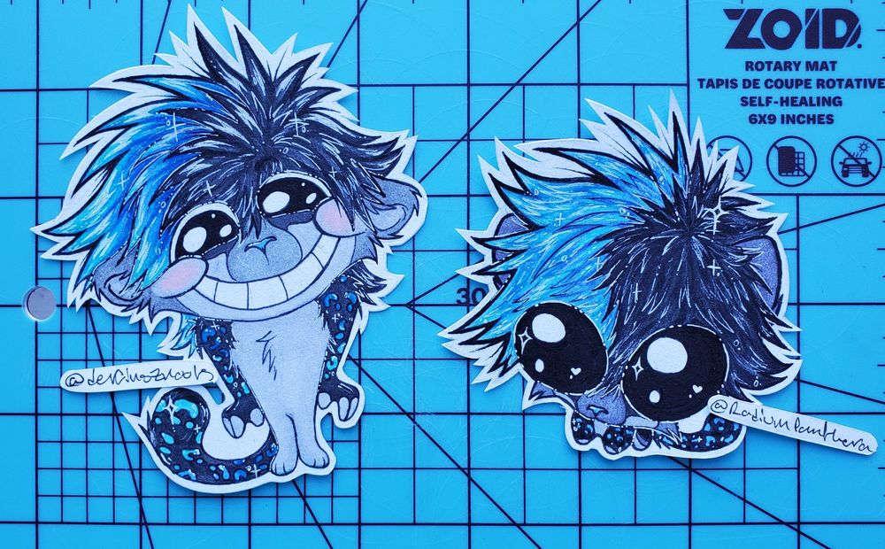 2 traditional pieces of my fursona Salem, stylized as discord emojis. on the left, he is standing upright with his arms tight to his body, his tail is curled upright on the viewers left, and he has big eyes and a toothy grin with blushy cheeks as if excited or very happy. His hair on the viewers left is dark to light blue, and the right half is black. There are lots of shines and sparkly stars in his hair and around his body. On the right, he is stylized as the autism creature, staring up at the viewer with a big head and big eyes, and little legs. He has the same hair as on the left, with heart and star sparkles in his eyes, hair, and some sparkles on his body as well. Both pieces are cut out paper on top of a blue cutting mat.