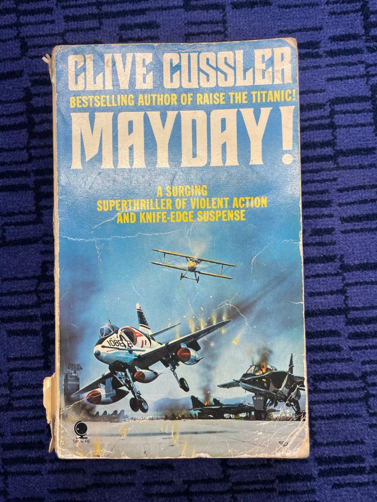 The lurid cover of Mayday! by Clive Cussler, featuring a burning fighter jet, a biplane, and the promise of 'a surging superthriller of violent action and knife-edge suspense'.