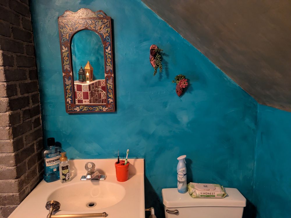 A photo of part of the wall in my bathroom that the vanity and toilet are up against. The wall is a fake lime wash look made with two different shades of aqua/ teal colored paint, the sloped ceiling has been painted metallic gold, there is a Moroccan hand-painted mirror hanging above the sink that has an arched top and is painted brownish red, green, off-white, gold, + an aqua that goes well with the wall color with a Moroccan floral/ leaf motif. There's a bottle of Listerine, hand soap, and a red orange toothbrush cup sitting on the vanity along with a brass hand towel bar resting on the edge of the sink. You can see that I have hung a small Moroccan babouche terracotta slipper that is pink and has trailing fake succulents coming out of it next to the mirror and following the angle of the ceiling, there is another more orange slipper with blue and black and red detailing hanging lower, and further above the toilet. You can see colorful brass and colored glass lanterns sitting on top of the bath surround in the reflection in the mirror. The bath surround is checkerboarded brownish red and Moroccan patterned tiles.