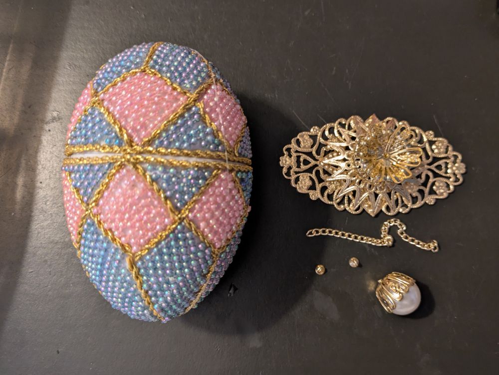 A photo of the previously described egg. Now both halves have been rubber banded together and you can't see the hinge but it is glued on on the back side here. Also, pictured are the stand, the finial that goes on top and the two little gold eyes and short length of gold tone chain that need to be installed last to keep the lid from flopping too far back.