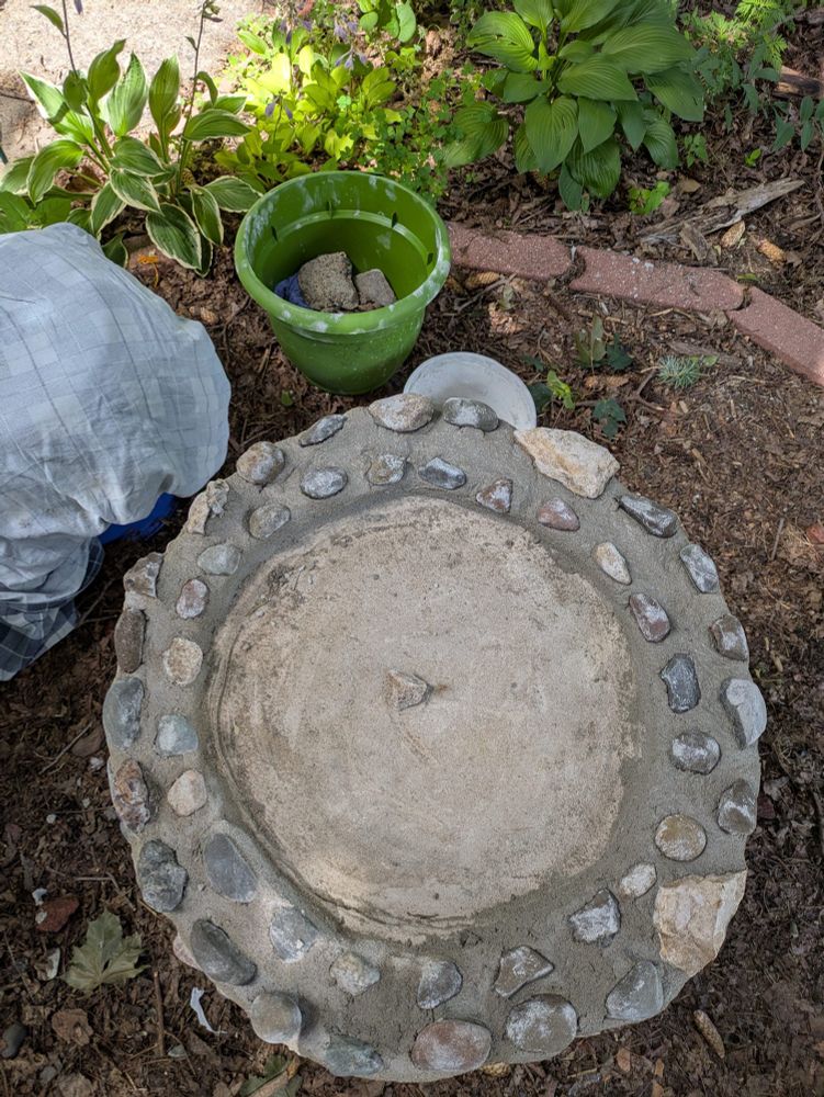 A photo of my little outside bird bath workstation showing the top of the bird bath with the first round of the edge wall complete, most of it still has wet mortar. You can also see a bright green planter filled with rocks, and a bucket being used as a stool with an inside out flannel bed sheet folded up on it as a cushion. You can see a couple hostas etc. In my garden behind this little tableau