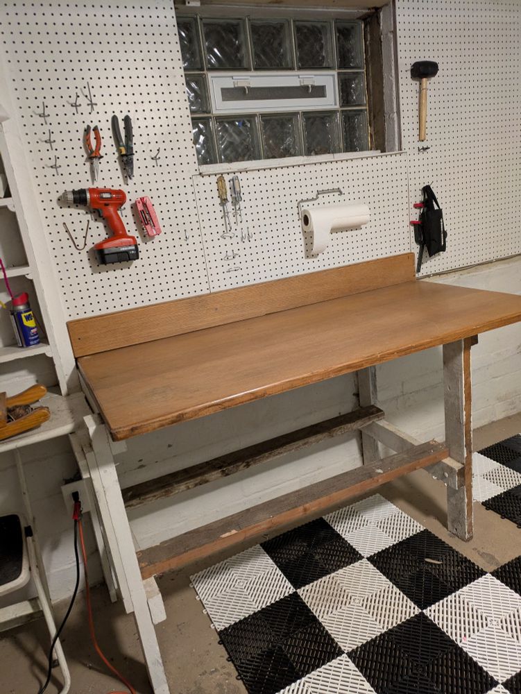 A photo of part of my basement workshop with the new upcycled desktop workbench on the original legs that were built for the old workbench. There are several tools kind of haphazardly and temporarily put on the pegboard above. The workbench and black and white checkerboard drainage flooring tiles need to be replaced and refigured.