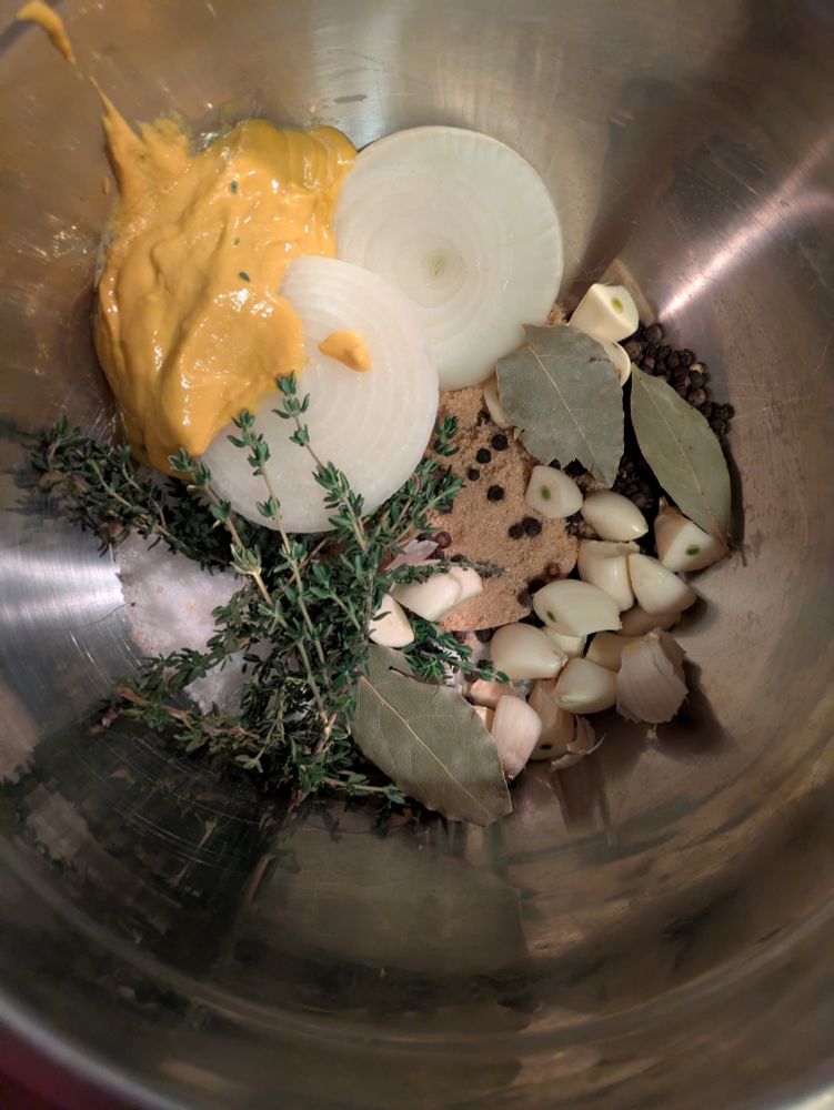 A large stainless steel bowl with a pile of ingredients in it, including a large white onion cut in half, mustard, several small sprigs of fresh thyme, three bay leaves, black peppercorns, brown sugar, and just a whole handful of garlic cloves cut in half