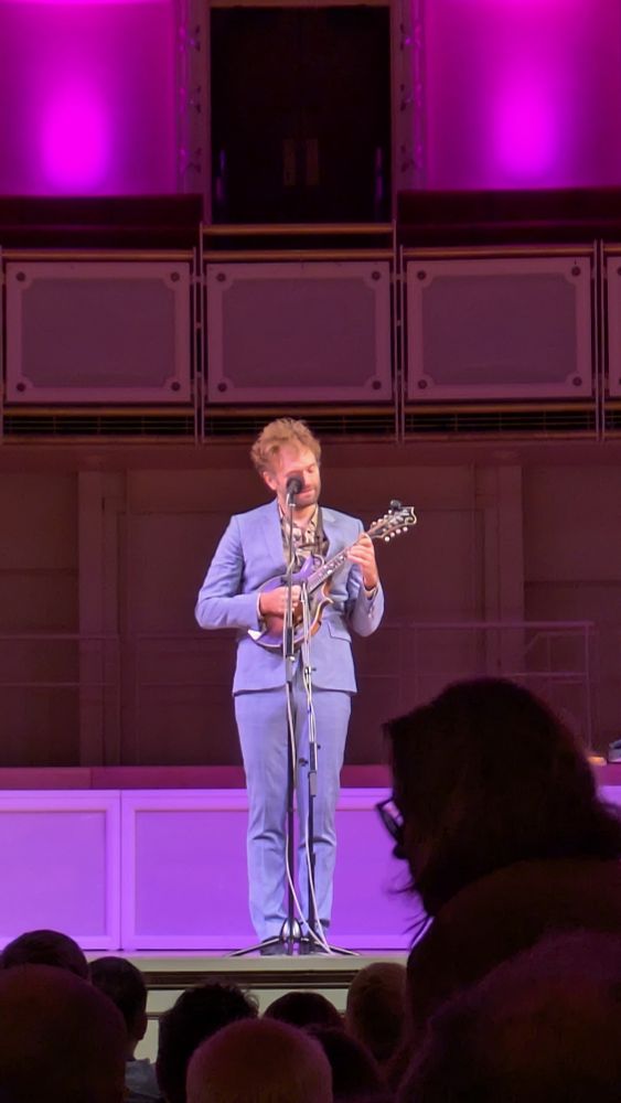 A man in a blue suit with wild hair stands on stage and plays a mandolin
