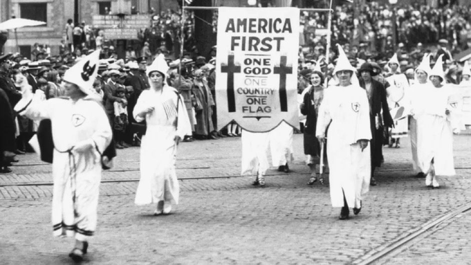 KKK parade black-and-white photo a long time ago White hoods and robes and the sign that says, America First.”