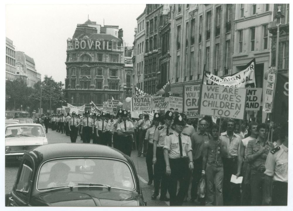 black and white photo 1974 pride march in London street men marching surrounded by police banners read we are natures children too 